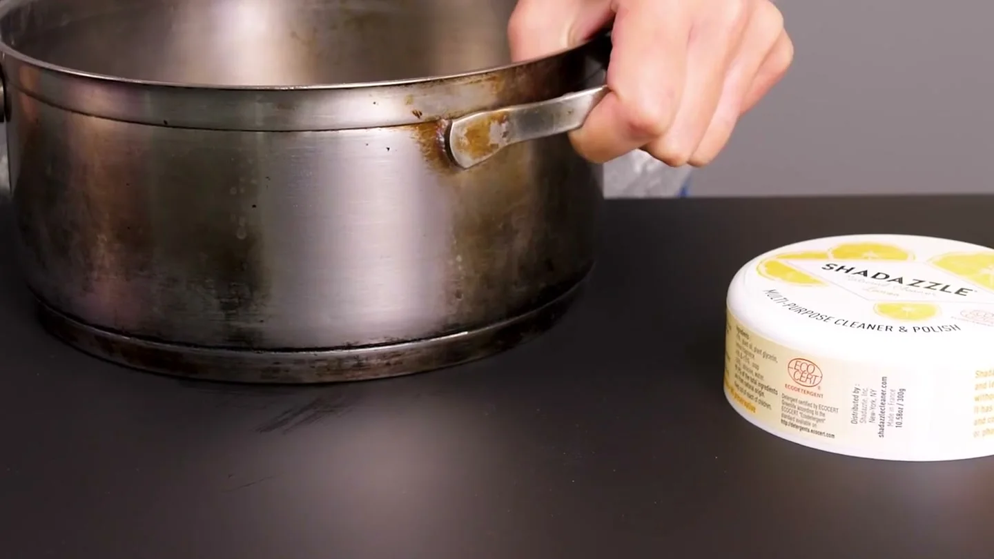 A clean stainless steel pot on a kitchen counter with a tub of lemon cleaning paste beside it